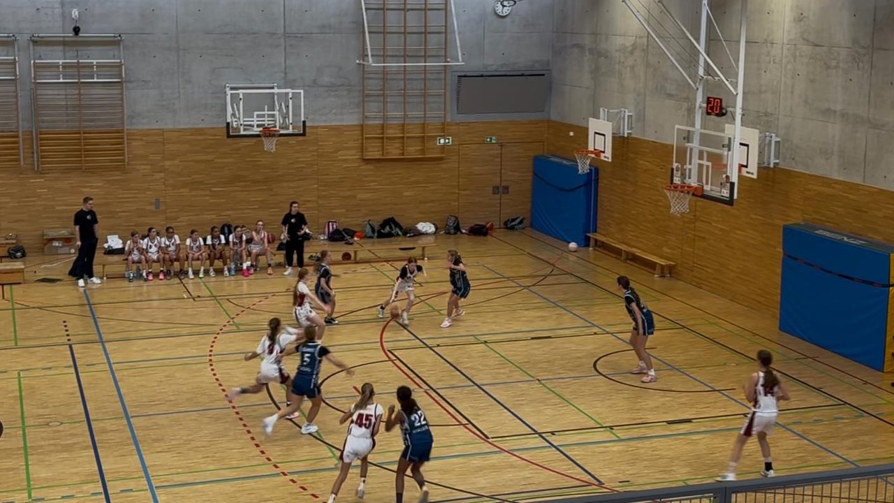 Spielszene in der Halle: Eine RegioTeam-Spielerin im weißen Trikot dribbelt, Spielerinnen der Basket-Girls in Blau verteidigen, im Hintergrund die Bank